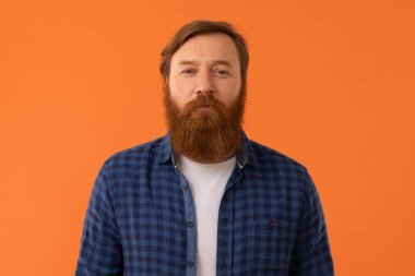 Kareli tişörtlü kızıl saçlı adam turuncu stüdyo arka planında sakin bir ifadeyle kameraya bakıyor. Ciddi bir adamın portresi, günlük kıyafetli kızıl saçlı.