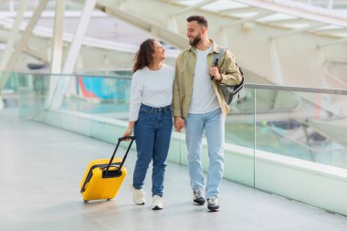 Genç bir çift, günümüzün cam duvarlı havaalanı terminalinde, seyahat ve birlikteliği simgeleyen sarı bir bavulla gelişigüzel yürüyor.