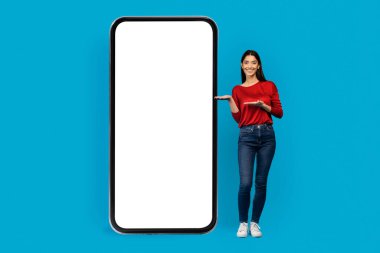 Günlük kıyafetlerle gülümseyen genç bir kadın boş akıllı telefon ekran modeli sunuyor, dijital uygulama ekranı için ideal, canlı mavi stüdyo arka planına karşı duran kadın, kolaj