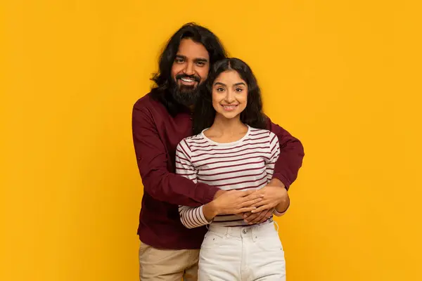 Man with arms around a woman, both smiling warmly on a solid yellow backdrop