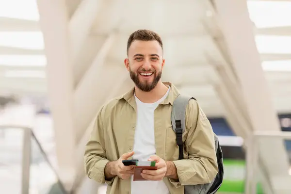 スマートフォンを持っている幸せな髭の男は楽しい表情で現代の空港に立っています — ストック写真