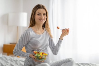 Neşeli hamile bir kadın renkli bir salata yer ve hamilelik süresince dengeli beslenmenin önemini vurgular.