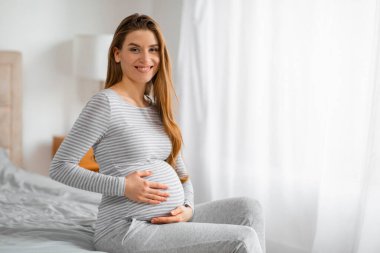 Hamile kadın rahat bir şekilde yatakta oturuyor, elleri karnında, hoş bir gülümseme takınıyor.