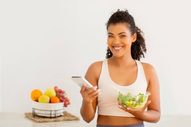 Mutlu bir milenyum kadını, vejetaryen yemek tarifi uygulamasını mutfaktaki akıllı telefonuyla gezer, spor kıyafetleri içinde durur, sağlıklı yaşam tarzını salatayla kucaklar, fitness ve beslenme alışkanlıklarını vurgular.