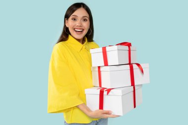 Şık sarı tişörtlü, şık, şık, genç esmer bir kadın. Elinde koyu kırmızı kurdeleli, şık beyaz hediyeler var. Mavi arka planda poz veriyor. Kutlama ve tatil kavramı.