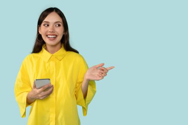 Özel bir teklif. Açık mavi stüdyo arka planında izole edilmiş cep telefonuyla ücretsiz fotokopi alanını gösteren heyecanlı genç bayan portresi. Pozitif kadın reklam için boş yer gösteriyor