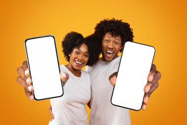 Binlerce yıllık heyecanlı Afro-Amerikan çifti beyaz ekranlı akıllı telefonları gösteriyor. Mobil uygulama reklamları için fotokopi alanı, online teklif, sarı stüdyo arka planında izole edilmiş.