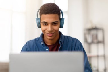 Modern kulaklık takan gülümseyen genç adam dizüstü bilgisayarın önünde oturuyor, evdeki ışık odasında çevrimiçi öğrenme seansına katılıyor.