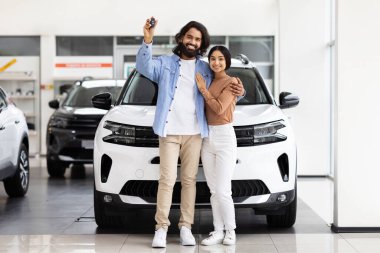 Bir araba galerisinde yeni bir beyaz aracın önünde kucaklaşan ve araba anahtarı tutan mutlu doğu çifti.