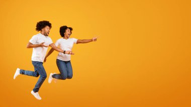 Heyecanlı Afro-Amerikan çifti, reklam için fotokopi çekilen uzay modelini işaret ediyor ve sarı arka planda izole edilmiş bir şekilde havaya zıplıyor. Siyah eşler harika bir aile teklifi sunuyor.
