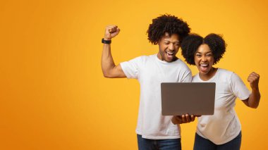 Duygusal genç siyah çift dizüstü bilgisayar ekranına bakıyor, yumruklarını sallıyor ve bağırıyor, Afrikalı Amerikalı online ticarette başarıyı kutluyor, sarı arka plan, kopyalama alanı