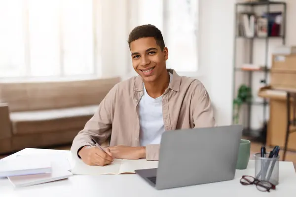 穿着休闲装的快乐的黑人男生一边在笔记本电脑上工作 一边在笔记本上记笔记 表达了他们的生产力感和满足感 — 图库照片