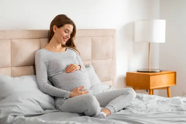 穏やかな妊娠中の女性はベッドの上に快適に座り 明るい寝室で腹を抱え 休息の瞬間を示唆し 彼女の胎児と結びついています — ストック写真