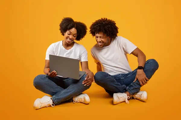 Pretty young black woman showing her husband something on laptop. Happy loving african american couple sitting on floor and using computer, checking educational website, yellow background
