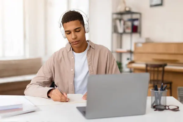 ヘッドフォンを身に着けている濃縮された黒人男性の学生は机に座り 自宅の明るい部屋のラップトップスクリーンからノートブックをコピーするためにメモを取ります — ストック写真