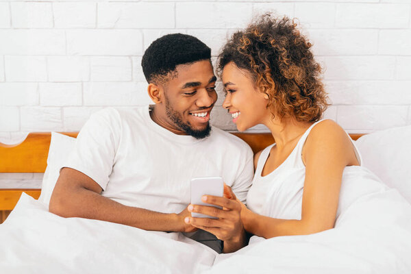 Между нами нет секретов. Счастливые афро-американец мужчина и женщина читают электронную почту по телефону, улыбаясь друг другу в постели
