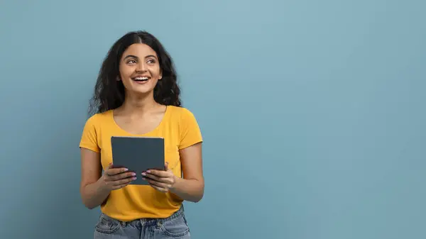 黄色いシャツを着た幸せな若い女性がデジタルタブレットを持ち 青い背景に楽しい表情を見つめています — ストック写真