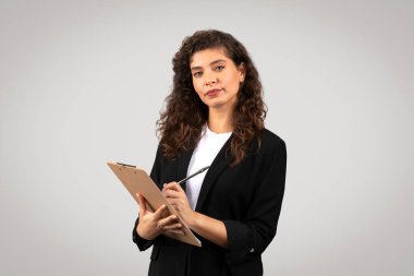 Curly-haired businesswoman holding a clipboard and pen with a contemplative expression clipart