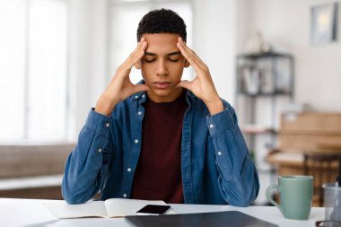 Genç bir adam başını tutarken rahatsızlığını ifade eder, muhtemelen dizüstü bilgisayarın önünde baş ağrısı ya da stres yaşar.