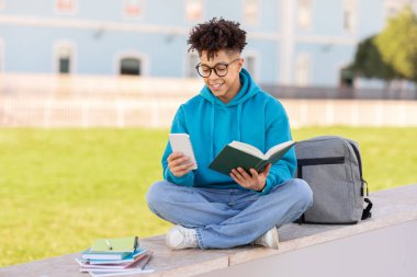 Odaklanmış Brezilyalı bir öğrenci kampüs bankında oturuyor, bir kitaba dalmış, yanında sırt çantası ve güneşli bir günde not defterleriyle.