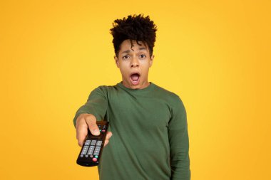 A surprised black man holding a remote control and expressing shock on a yellow background clipart