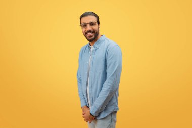 Indian man with a beard and glasses smiles while looking over his shoulder against a yellow background clipart