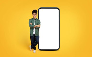 Confident african american man in casual clothes leaning on a gigantic smartphone mockup against a yellow backdrop clipart
