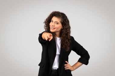 Confident young woman with curly hair pointing at the camera, wearing a blazer over a casual shirt clipart