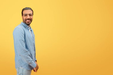 Confident and cheerful indian man with glasses and beard posing against a yellow background clipart