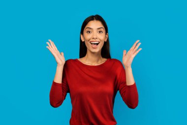 Şaşırmış genç bir kadın ellerini kaldırıyor ve heyecanla kameraya bakıyor, güzel bir teklif veya tanıtıma duygusal tepki veren, mavi arka planda tek başına duran,