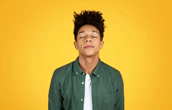 A young black man in a green shirt stands serenely with his eyes closed against a yellow background, exuding calmness