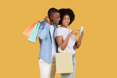 Neşeli genç Afro-Amerikan çifti alışveriş çılgınlığı içinde. Akıllı telefon ekranındaki bir buluş için heyecanlarını paylaşıyorlar. Sarı arka plandaki çantalar, stüdyo.