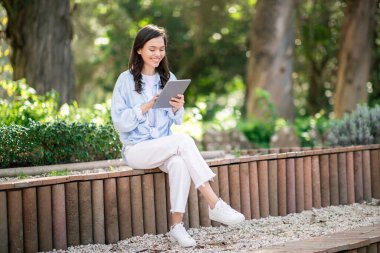 Avrupalı genç bir kız öğrenci parkta bir bankta bağdaş kurarak oturuyor, açık havada, barışçıl, yeşil bir arka planda tablet ekranına odaklanmış. Eğitim, eğitim.