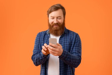 Yakışıklı kızıl sakallı adam stüdyo ortamında cep telefonuna bakarken gülümsüyor modern iletişimi somutlaştırıyor turuncu arka planda akıllı telefondan mesaj atıyor. Teknoloji ve aletler.