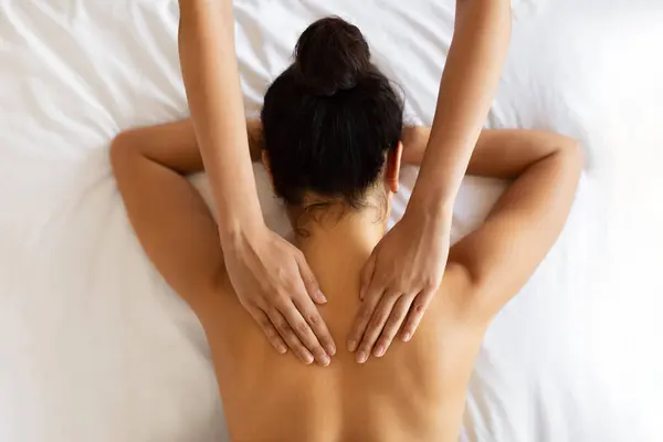 Above view of woman receiving healing body massage from unrecognizable therapist at luxury spa, lying on white bed, masseuses hands on female clients back. Top view shot