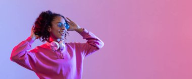 Pembe kapüşonlu neşeli genç kadın, güneş gözlüğü takıyor ve beyaz kulaklıkla müzik dinliyor. Renkli pembe ve mavi arka plan, panorama, serbest alan...