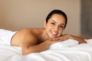 Çekici, esmer, milenyum kadını, kameraya gülümseyen, kapalı yatakta dinlenen, modern lüks spa salonundaki güzellik tedavi seansını beklerken dinlenen.