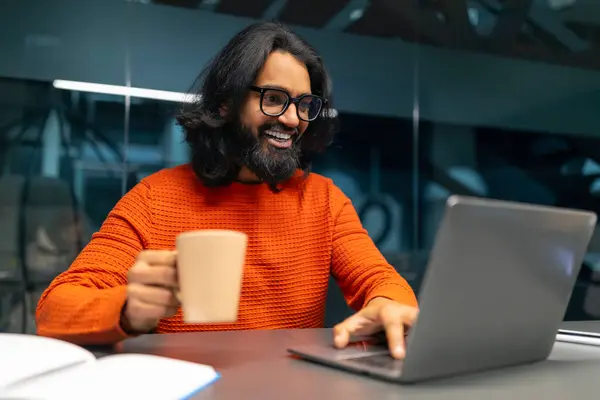 メガネとオレンジのセーターを着ている男性は ラップトップを使用しながらコーヒーマグを保持し テーブルの上の本 — ストック写真