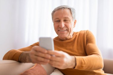 Son sınıf öğrencisi rahat bir erkek oturuyor ve akıllı telefonuyla meşgul. Ev ortamında hoş bir yüz ifadesi sergiliyor.