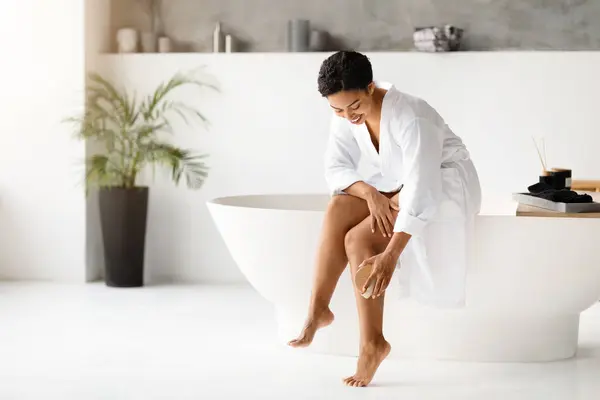 在家里 身穿浴衣干洗腿的黑人妇女微笑着 非洲裔美国妇女在独立的浴缸和装饰过的植物的宁静浴室里操练皮肤剥脱 — 图库照片