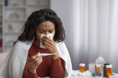 Hasta siyah kadın akan burnu temizliyor ve evde termometreye bakıyor, Afrika kökenli Amerikalı kadın oturma odasındaki kanepenin üzerinde battaniyeyle otururken sıcaklığı kontrol ediyor.