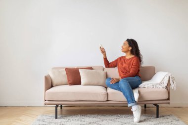 Siyah Genç Kadın Uzaktan Kumanda ile Evde Klima Açıyor, Oturma odasında koltukta dinlenirken Mutlu Afrikalı Amerikalı Kadın Sıcaklığı Ayarlıyor, Boşluğu Kopyala