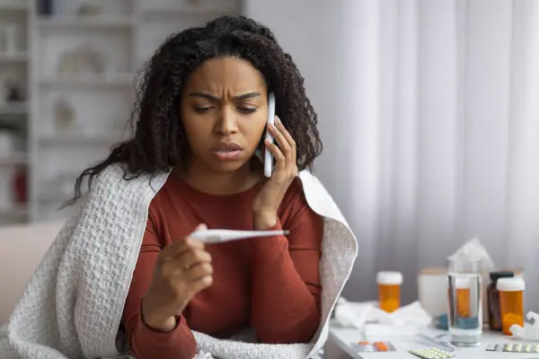 网上咨询 生病的黑人妇女拿着温度计 一边坐在沙发上一边用手机叫医生 一边给生病的非洲裔美国妇女放空 — 图库照片