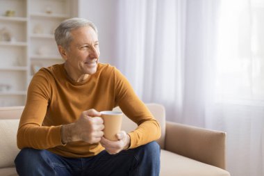 Yaşlı bir adam koltukta rahat bir şekilde oturur, sıcak bir fincan tutar, rahatlamayı ya da kahve molasını gösterir.