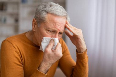 Hasta, gri saçlı yaşlı bir adam hapşırıyor ve başına dokunuyor, sağlıksız hissediyor. Büyükbabam soğuk algınlığı ya da gripten muzdarip, ateşi var, yakın plan. Yaşlılar sağlık konsepti