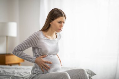 Braxton Hicks contractions. Young pregnant female feeling pain, suffering from abdominal ache at home, worried millennial woman sitting on bed and touching her big tummy, having cramp, copy space clipart