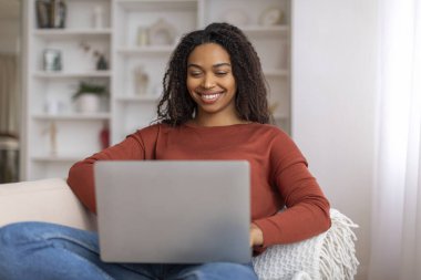 Rahat siyahi kadın evde çalışmaktan zevk alıyor. Oturma odasındaki rahat koltukta otururken dizüstü bilgisayarını kullanıyor. Mutlu Afro-Amerikan kadın internet işi yapıyor.