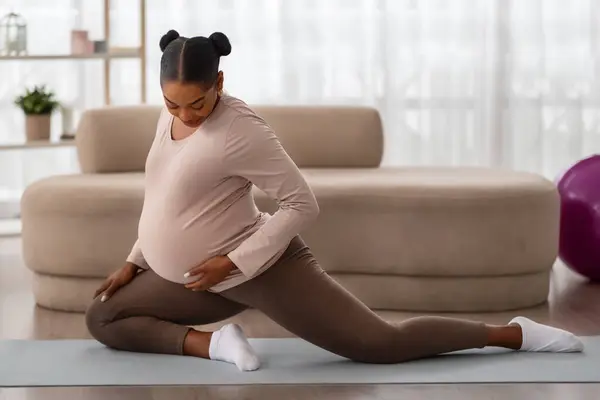 アクティブ妊娠コンセプト 笑顔の黒人女性将来のママは彼女の足を伸ばし 自宅のジムでマットで朝ヨガのトレーニングを行っています 国内のフィットネスを行っているかなり期待される女性 屋内で運動 — ストック写真