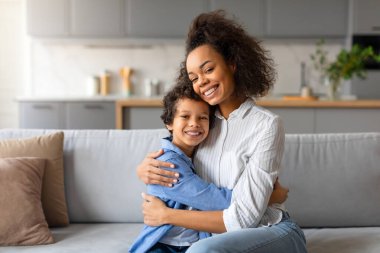 A loving African American mother embraces her young son in a cozy home setting clipart