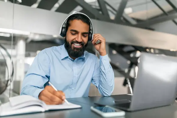 Smiling bearded millennial indian guy employee have online training, sitting at desk at modern office, using headset and laptop computer, taking notes. Professional education concept